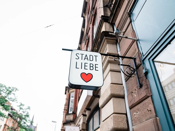 Stadtliebe_Vorderer Westen Das Schild vom Geschäft Stadtliebe ist Sichtbar. Auf dem Schild ist unter der Schrift ein rotes Herz abgebildet. Im Hintergrund kann man den Stadtteil Vorderer Westen in Kassel erkennen.