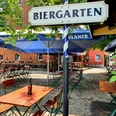 Biergarten mit Holztischen und -stühlen, umgeben von grünen Bäumen, unter blau-weißen Schirmen.