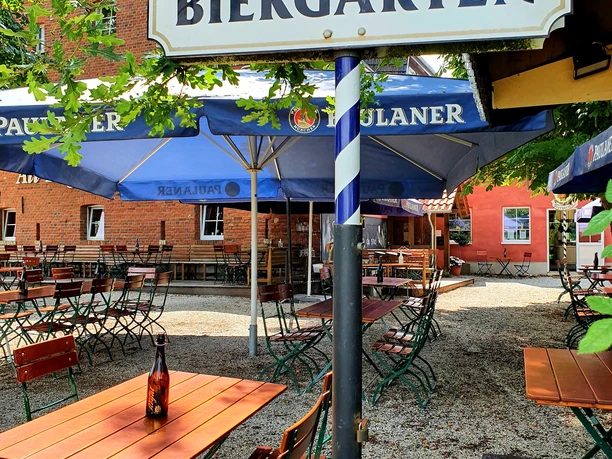 Biergarten mit Holztischen und -stühlen, umgeben von grünen Bäumen, unter blau-weißen Schirmen.