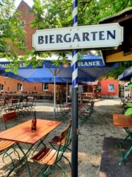 Alt-Enginger Mühle: Biergarten Biergarten mit Holztischen und -stühlen, umgeben von grünen Bäumen, unter blau-weißen Schirmen.