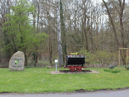 Ein blumenbepflanzter Lorewagen steht neben einem Gedenkstein in einer grünen Landschaft am Waldrand.