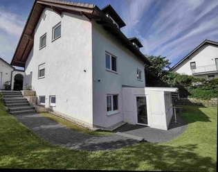 Fewo Orchidee.jpg Weißes zweistöckiges Wohnhaus mit Erker und Schindeldach, umgeben von gepflegtem Rasen und Pflanzen.