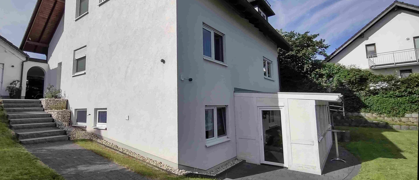 Fewo Orchidee.jpg Weißes zweistöckiges Wohnhaus mit Erker und Schindeldach, umgeben von gepflegtem Rasen und Pflanzen.