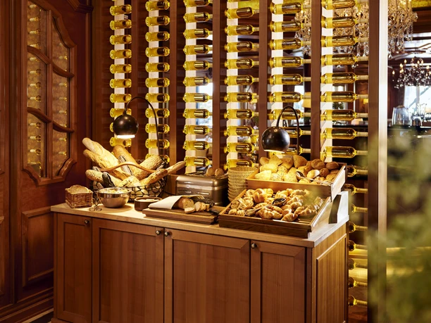 Guetersloh-Parkhotel-Frühstück Parkhotel_15Teutoburger-Wald-©-Viveno-Group-GmbH-Studio-Röcken.jpg Holzverkleideter Raum mit Regal voller Weinflaschen, vorn Korb mit Brot und frischen Backwaren.