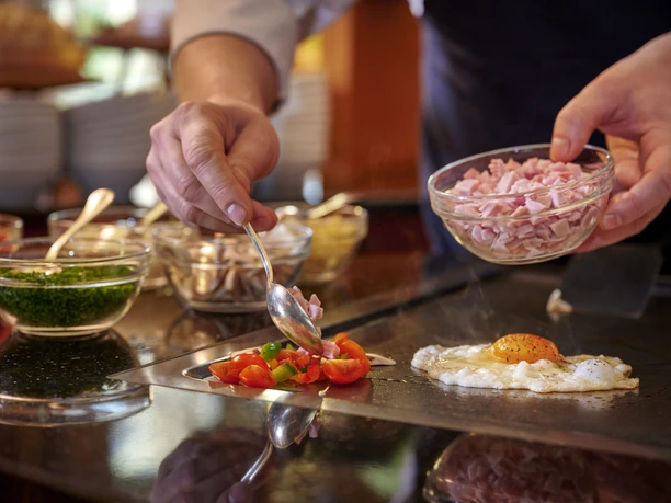 Guetersloh-Parkhotel-Frühstück Parkhotel_09Teutoburger-Wald-©-Viveno-Group-GmbH-Studio-Röcken.jpg Ein Koch bereitet ein frisches Spiegelei zu, umgeben von Gemüse, Kräutern und fein geschnittenem Schinken.