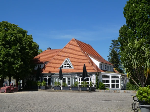 Backsteinbau mit rotem Ziegeldach, außen weiße Fenster, umgeben von Bäumen und einem gepflasterten Platz.