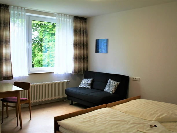 Helles Doppelzimmer mit Sofa und Fenster, schlicht eingerichtet, Blick auf grüne Außenlandschaft.