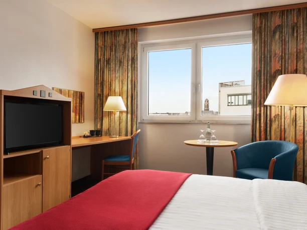 Hotelzimmer mit Doppelbett, roter Decke, Schreibtisch, Fernseher und Fenster mit Stadtblick.