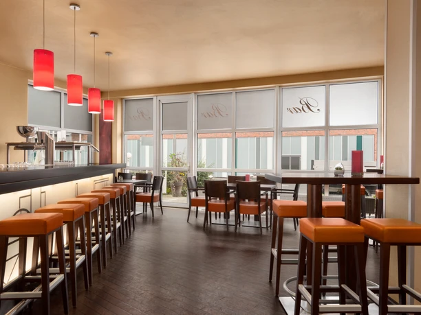 Helle Hotelbar mit hohen Fenstern, roten Pendelleuchten und orange gepolsterten Barhockern.