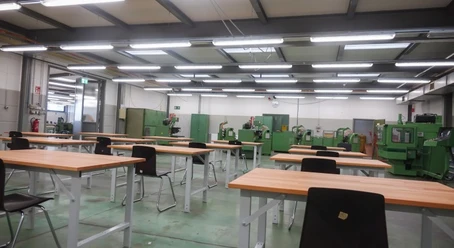Lernwerkstatt.jpg Heller Raum mit Werkbänken, Stühlen und grünen Industriemaschinen im Hintergrund.