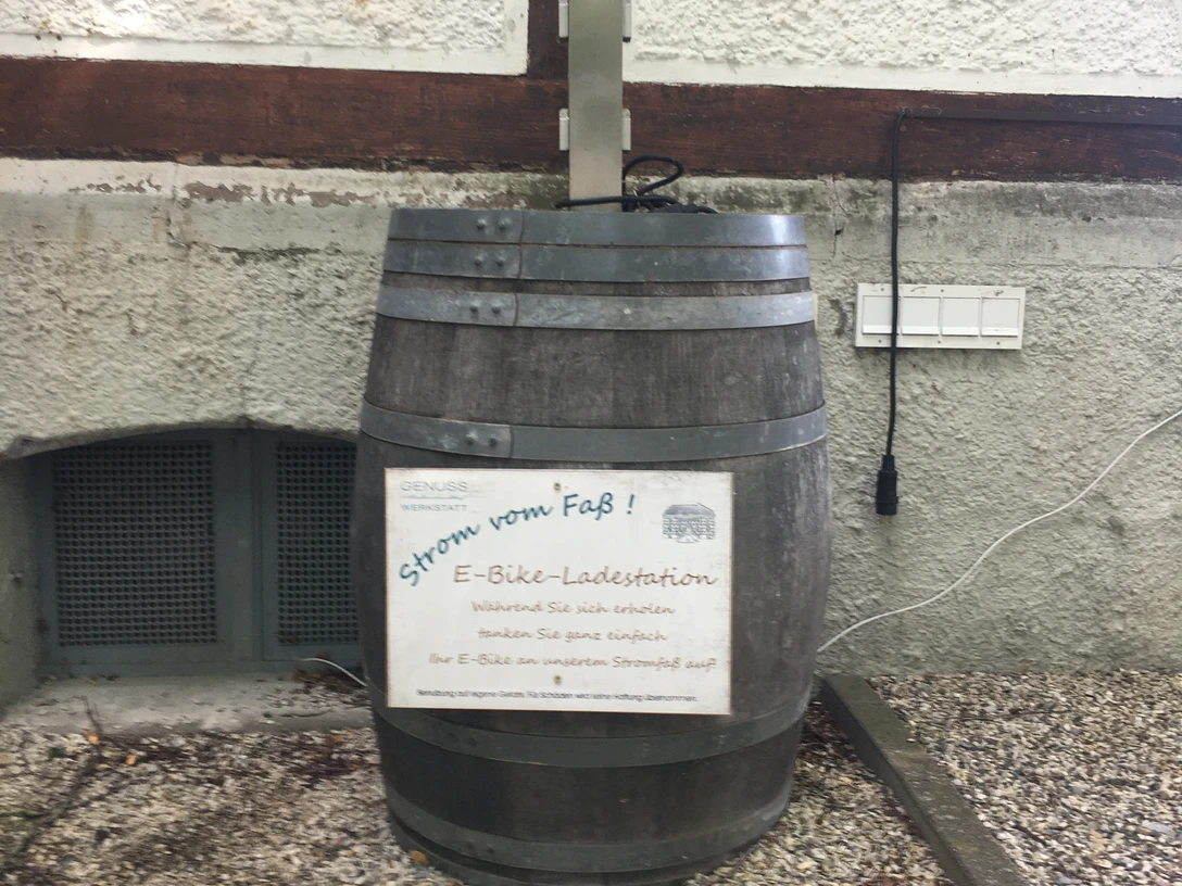 E-Bike-Ladestation in einem hölzernen Fass integriert, neben einer Hauswand auf Kiesuntergrund.
