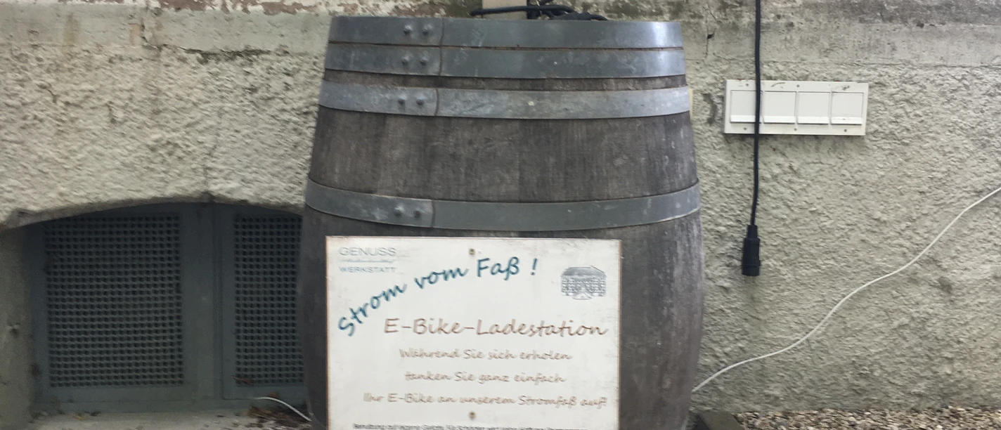 E-Bike-Ladestation am Mallinckrodthof.JPG E-Bike-Ladestation in einem hölzernen Fass integriert, neben einer Hauswand auf Kiesuntergrund.