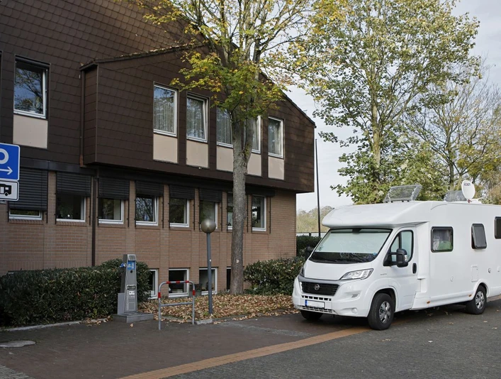 Stellplatz für Wohnmobile neben einem Gebäude mit Bäumen und Parkautomat, herbstliche Umgebung.