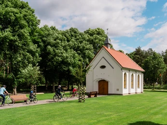 Weiße Kapelle mit rotem Dach, umgeben von grüner Wiese und Bäumen, vor der Radfahrer vorbeifahren.