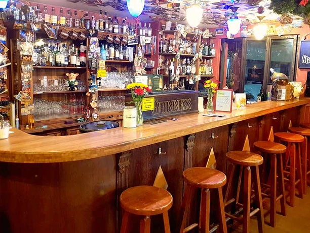 cottage-bottrop-5.jpg Holztheke mit Barhockern in einem Pub, dahinter Regale voller Gläser, Flaschen und bunter Dekoration.