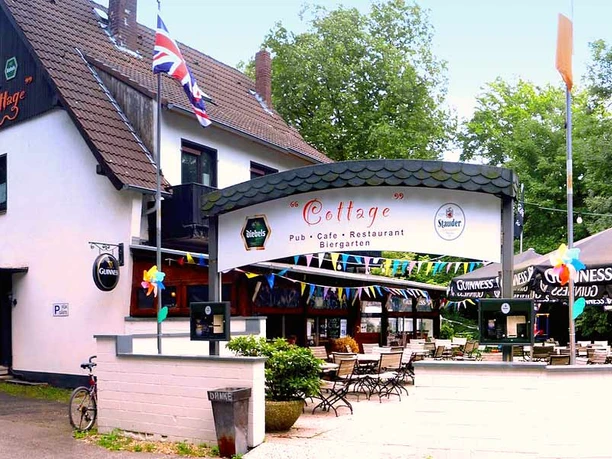 cottage-bottrop-6.jpg Eingangsbereich des Cottage Pub in Bottrop mit Biergarten, bunten Fahnen und Außensitzplätzen.