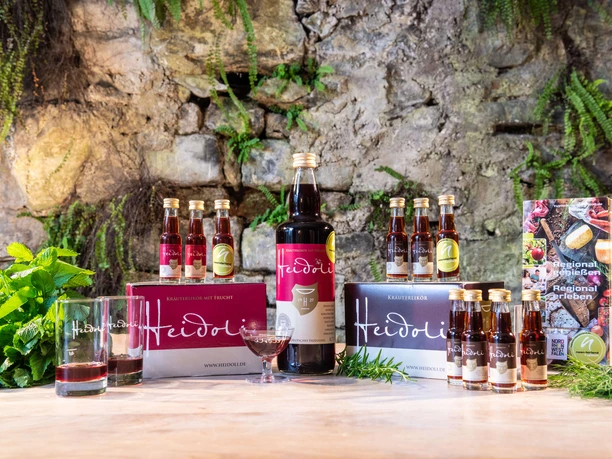 Heidoli - Getränke Doppstadt GmbH Eine Auswahl von Heidoli Getränken mit befüllten Gläsern und einem Prospekt, dekoriert auf Holz vor Steinwand.