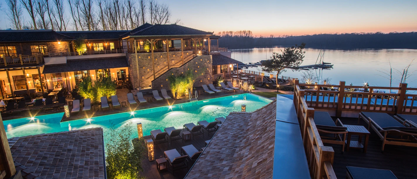 Vabali Spa in Hilden Ein modernes Spa mit Außenpool, umgeben von üppigem Grün und entspannender Flusslandschaft in der Abenddämmerung.