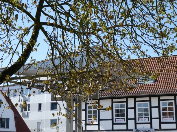 The carillon in Achim is located under a tree and is surrounded by half-timbered buildings.