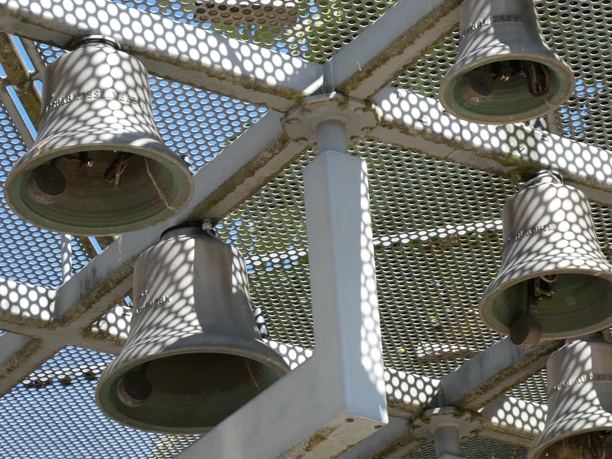Sechs große, teils schattige Glocken hängen in einem Metallgerüst gegen den blauen Himmel.