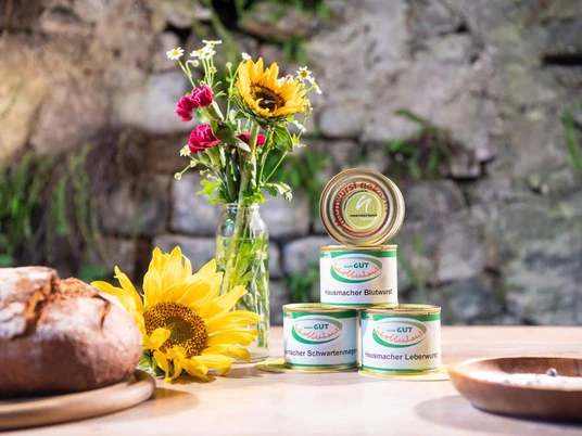 Gut Schobbenhaus aus Mettmann Verschiedene Dosen Wurst und frische Sonnenblumen auf einem rustikalen Holztisch vor einer Steinwand.