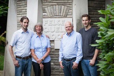 Familie Römer der Haaner Felsenquelle Eine Familie steht vor einem Steinmonument mit der Aufschrift "Felsenquelle 1903". Alle vier Personen lächeln in die Kamera und tragen lässige Kleidung. Im Hintergrund sind steinerne Strukturen und grünes Laub zu sehen.