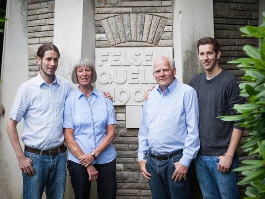 Familie Römer der Haaner Felsenquelle Eine Familie steht vor einem Steinmonument mit der Aufschrift "Felsenquelle 1903". Alle vier Personen lächeln in die Kamera und tragen lässige Kleidung. Im Hintergrund sind steinerne Strukturen und grünes Laub zu sehen.