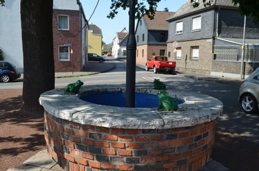 Kradepohl in Monheim am Rhein Ein runder Brunnen aus Ziegelsteinen mit Froschfiguren steht an einer Straßenkreuzung in Monheim am Rhein.