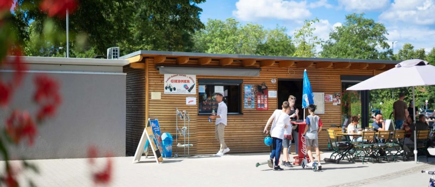 Tourist-Information Giebner in Monheim am Rhein Tourist-Information mit Holzverkleidung, Menschen und Fahrradführungen, umgeben von grünem Park.
