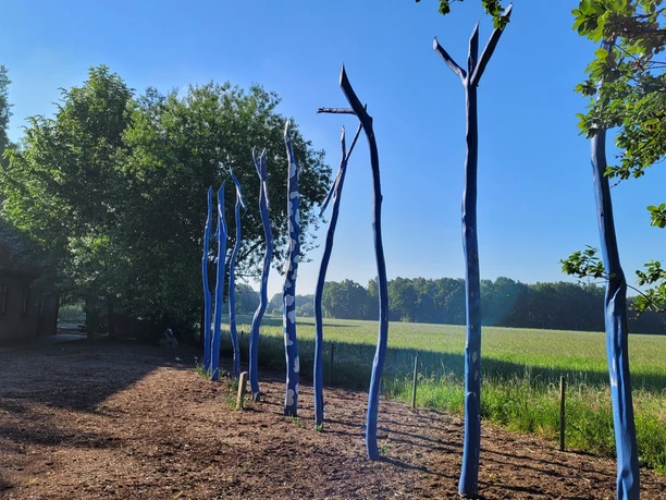 NRW Nordpunkt Sieben blau bemalte Holzstämme stehen auf einer Wiese, umgeben von Feldern und Bäumen im Sonnenschein.