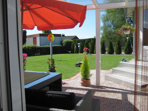 Sonnige Terrasse mit rotem Sonnenschirm, Blick auf gepflegten Garten und Rasen, moderne Sitzmöbel.