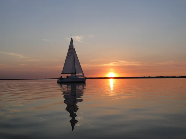 Sailing boat glides calmly over still water while the radiant sun sets on the horizon.