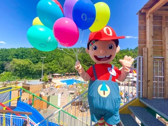 Maskottchen Oskar Oskarshausen: Ein fröhliches Maskottchen in rotem Outfit hält bunte Luftballons in einem Freizeitpark; im Hintergrund sind Bäume und Spielbereiche zu sehen.