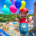Maskottchen Oskar  Oskarshausen: Ein fröhliches Maskottchen in rotem Outfit hält bunte Luftballons in einem Freizeitpark; im Hintergrund sind Bäume und Spielbereiche zu sehen.