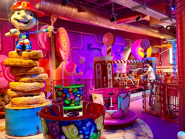 Oskarshausen: Colorful indoor children's attraction with giant lollipops, a cheerful figure on cookies and colorful teacups in a lively atmosphere.