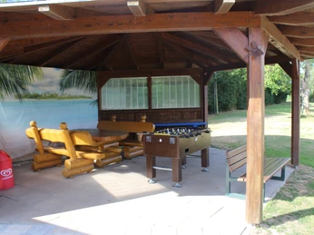 Überdachte Sitzgruppe mit Holzbänken und Tisch, Kicker, Mülleimer und Strandbild im Hintergrund.