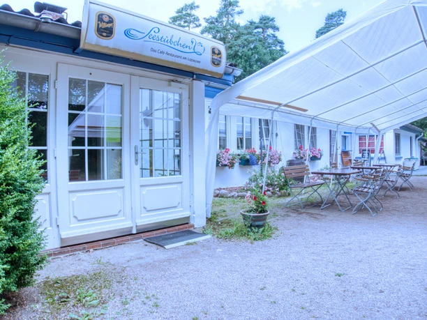 Außen.jpeg Weißes Gebäude des Seestübchens mit Eingang und überdachter Terrasse mit Tischen und Stühlen.