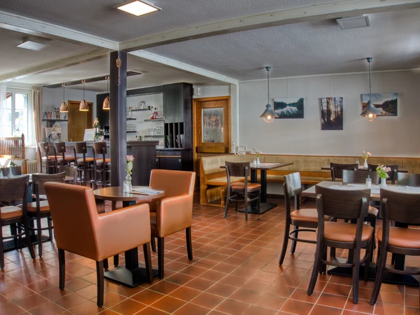 Café Heller Gastraum einer Pension mit Theke, Holztischen und braunen Sesseln auf Fliesenboden.