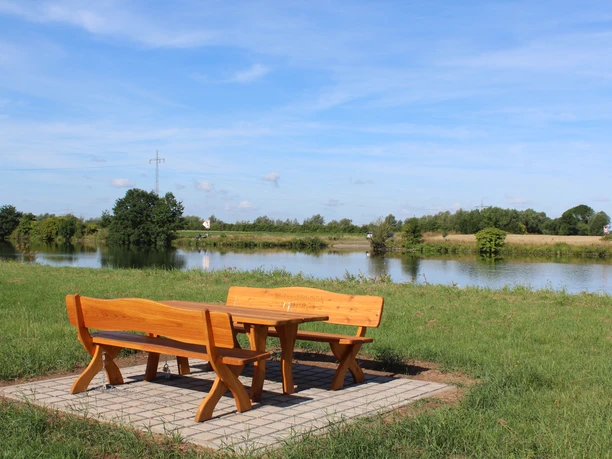 Holztische und Bänke auf gepflasteter Fläche mit Blick auf einen Teich und grüne Landschaft.