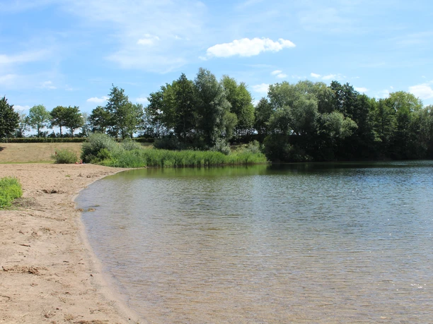 Badesee Lahde Ein ruhiger Badesee mit einladendem Sandstrand und üppigem grünen Uferbewuchs unter blauem Himmel.