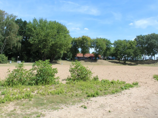 Badesee Lahde Sandstrand umgeben von grünen Bäumen, im Hintergrund sind Gebäude mit roten Dächern sichtbar.