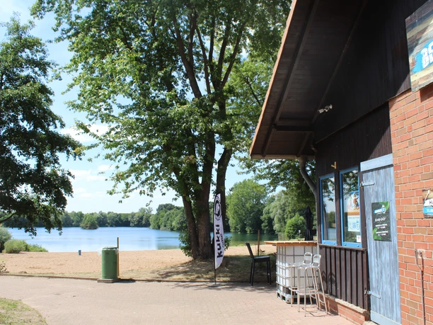 Badesee Lahde mit Sandstrand, im Vordergrund ein Kiosk; umgeben von Bäumen und ruhigem See.