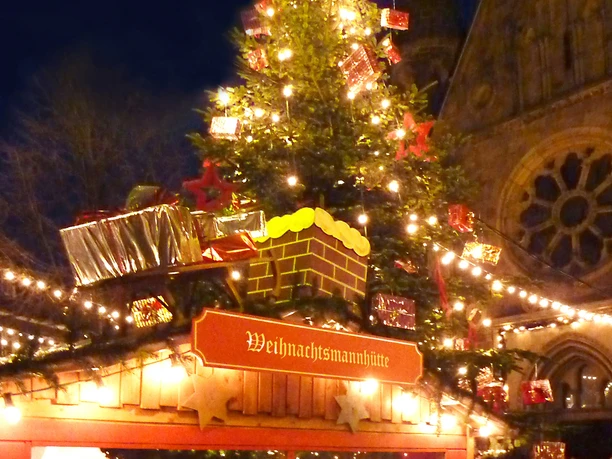 Blotschenmarkt Weihnachtsmannhütte mit Weihnachtsbaumund im Hintergrund Weihnachtsmannhütte mit festlich geschmücktem Baum, umgeben von Lichtern vor einer Kirche bei Nacht.