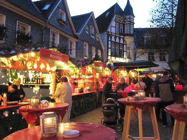Weihnachtsmarkt mit Fachwerkhäusern, beleuchteten Buden, Menschen beim Einkaufen und roten Tischdecken.