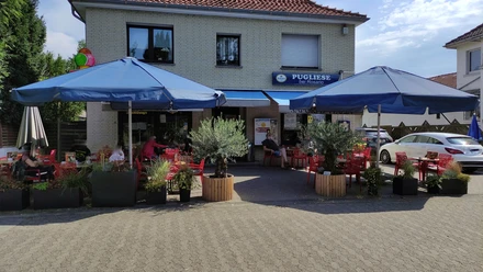 Eiscafé Pugliese.jpeg Eiscafé Pugliese mit blauen Sonnenschirmen, roten Tischen und Pflanzen auf der Terrasse an einem sonnigen Tag.
