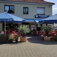 Eiscafé Pugliese.jpeg Eiscafé Pugliese mit blauen Sonnenschirmen, roten Tischen und Pflanzen auf der Terrasse an einem sonnigen Tag.