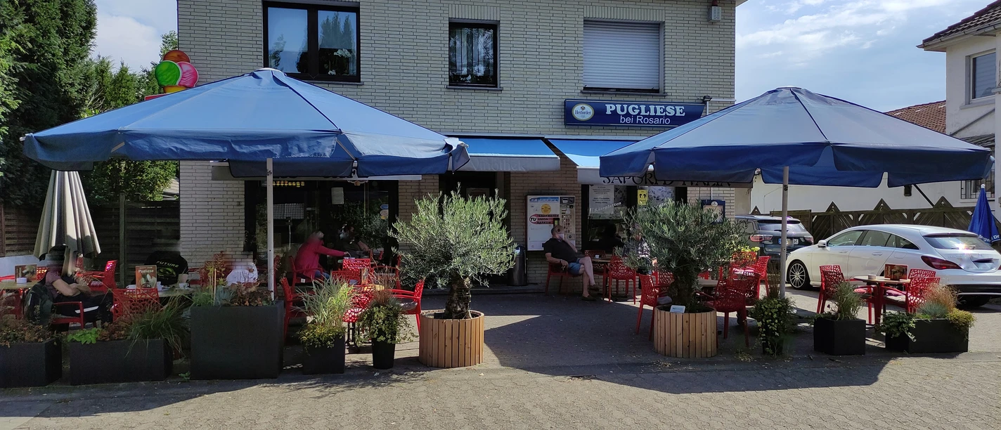 Eiscafé Pugliese.jpeg Eiscafé Pugliese mit blauen Sonnenschirmen, roten Tischen und Pflanzen auf der Terrasse an einem sonnigen Tag.