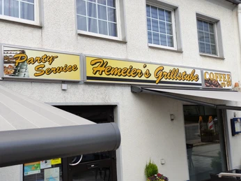 Schild von Hemeiers Grillstube über Eingangsbereich eines Gebäudes mit grauer Fassade und Markise.