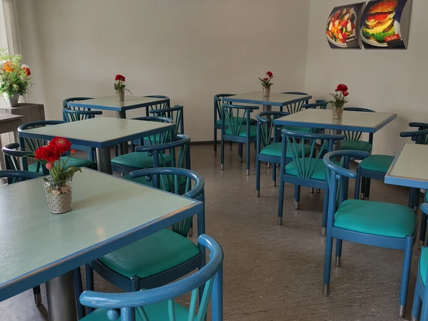 Mehrere blaugrüne Tische und Stühle in einem hellen Raum, rote Blumen dekorieren die Tische.