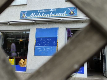 Äußere Vorderansicht des Geschäfts Mühlenbernd mit Werbung auf Schaufenstern sichtbar hinter einem Holzzaun.